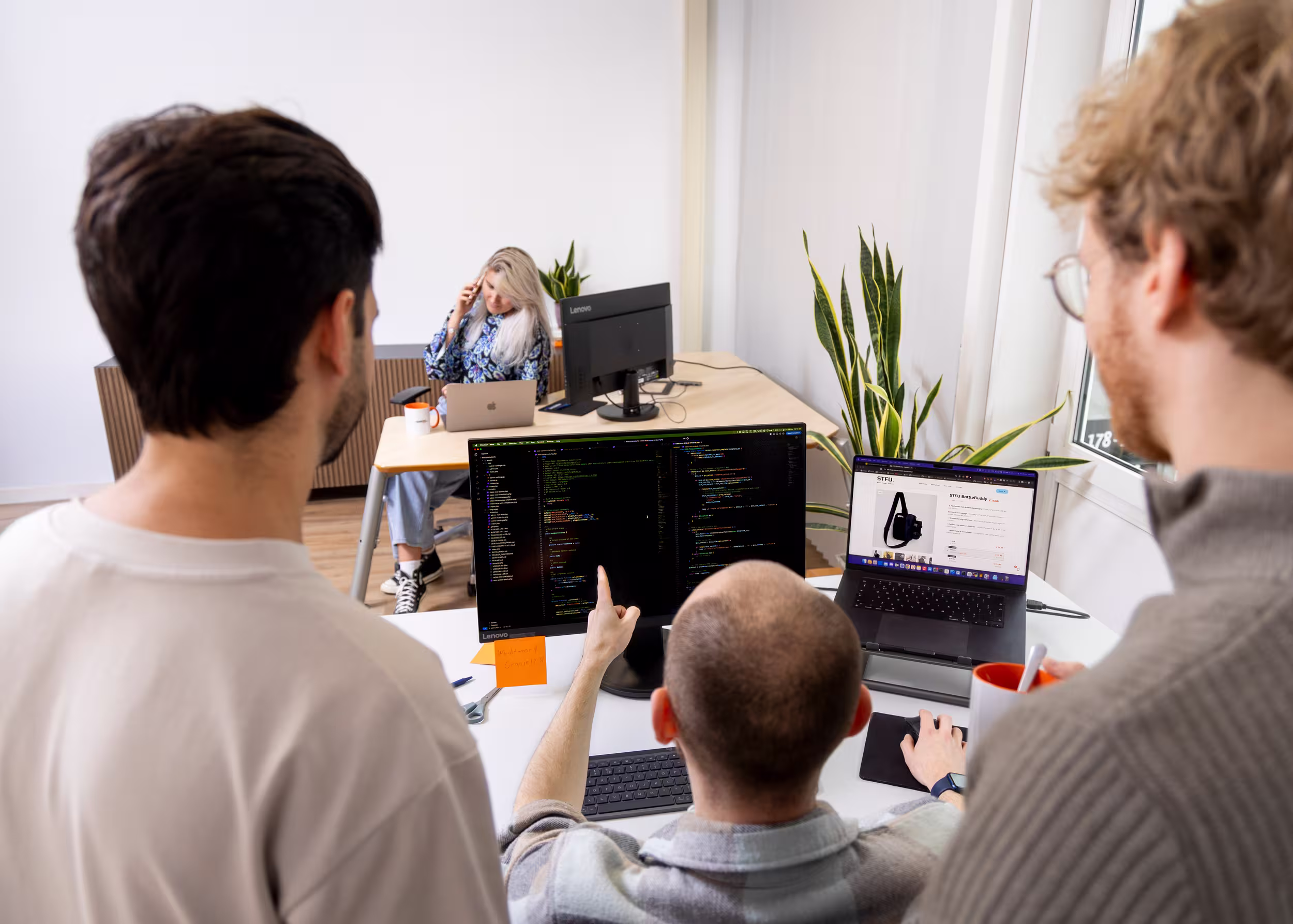 Collega's bespreken samen de code op een beeldscherm, op de achtergrond belt een collega