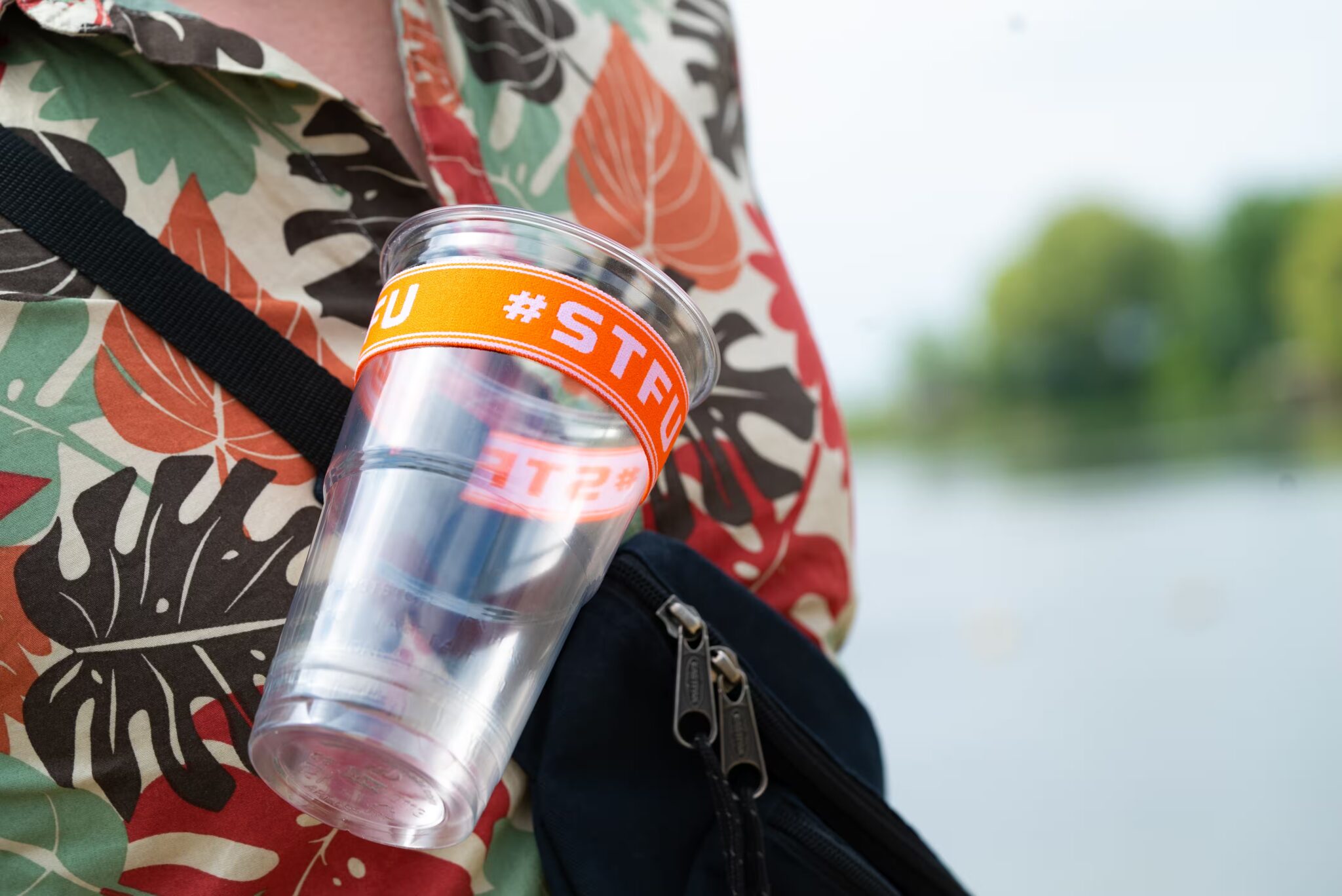 Doorzichtige festivalbeker met oranje STFU-band, vastgemaakt aan een rugzakriem